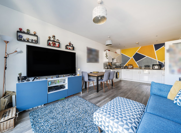 Living room with sofa, television, shelving and dining table, leading into kitchen.