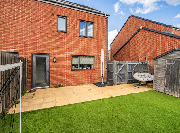 Fenced rear garden with lawn, patio, wooden shed and red brick house.