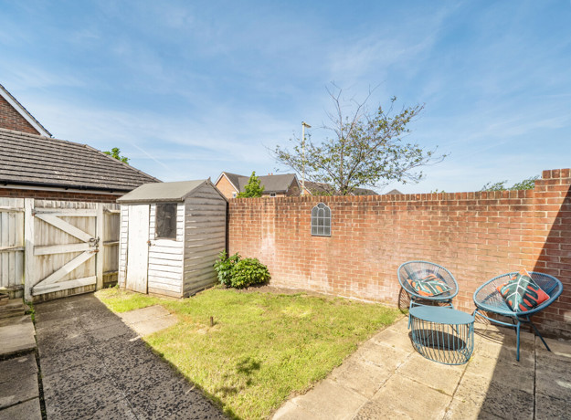 Backyard with paved area with small table and 2 chairs, grass patch, and garden shed.