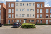 Apartment block with brick facade, cream panels, parked cars, and shrubs.