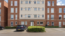 Apartment block with brick facade, cream panels, parked cars, and shrubs.