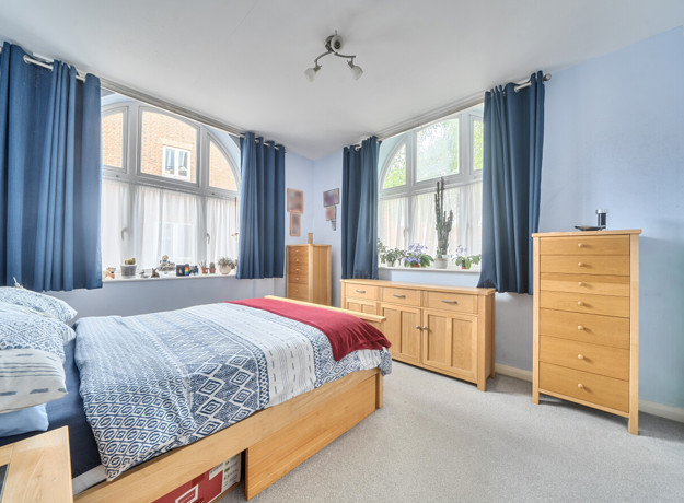 Bedroom with double bed, blue bedding, wooden dresser and cabinets with drawers, and large windows.