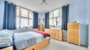 Bedroom with double bed, blue bedding, wooden dresser and cabinets with drawers, and large windows.