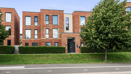 Modern brick apartment building with balconies, trees, and grass verge by the road.
