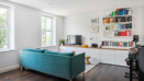 Living area with a teal sofa, wall-mounted shelves, piano and windows.