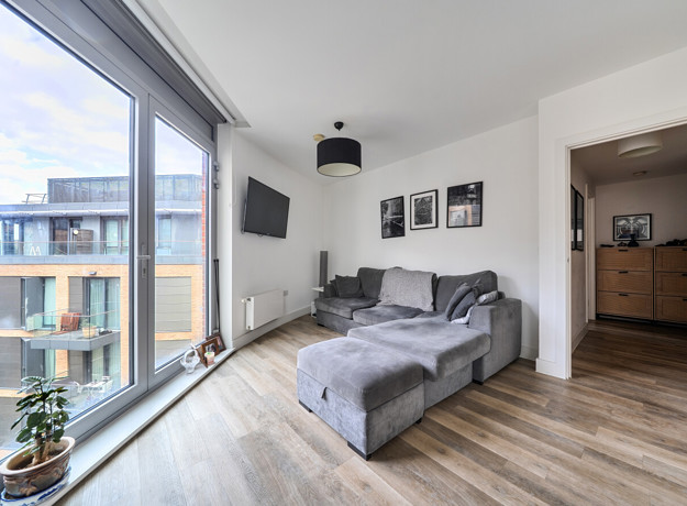 Living space with wall art, sofa beside floor-to-ceiling windows and a door leading into a hall.