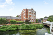 Brick building beside a canal with greenery and a nearby bridge.