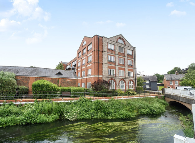 Brick building beside a canal with greenery and a nearby bridge.
